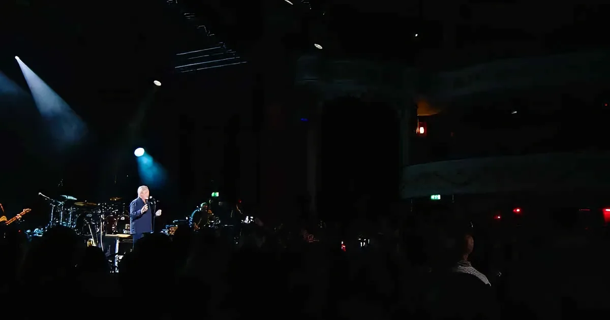 A man in a dark suit stands on stage, illuminated by a spotlight, with musicians and equipment behind him.