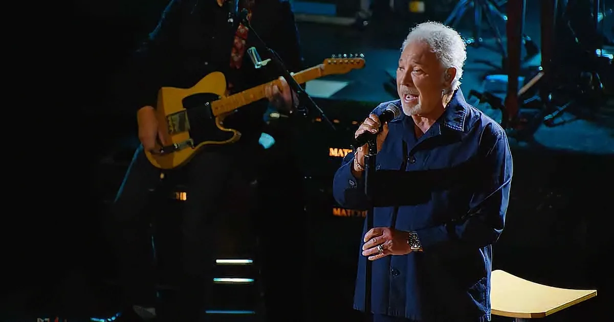 An older man in a blue shirt sings into a microphone on stage, with a guitarist in the background.