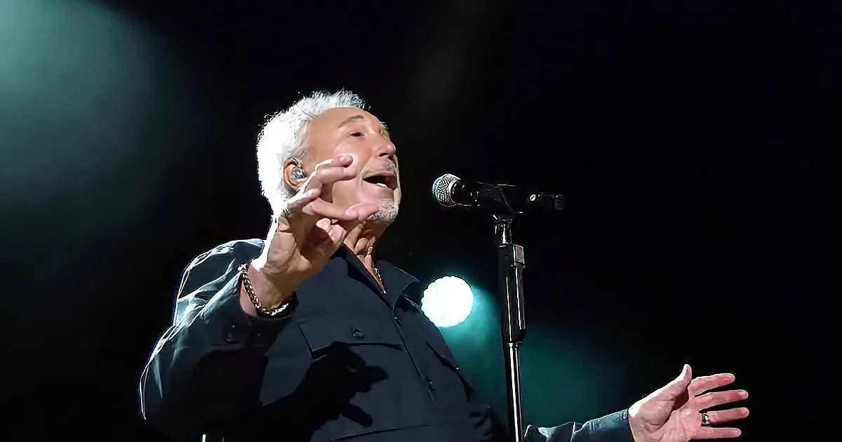 An older man with white hair and a black shirt is singing into a microphone on stage.