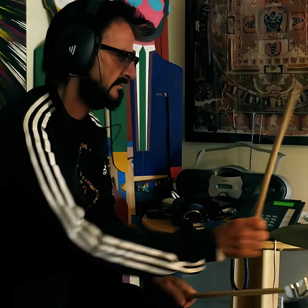 A man wearing headphones and a black Adidas tracksuit holds drumsticks, surrounded by colorful artwork and musical equipment.