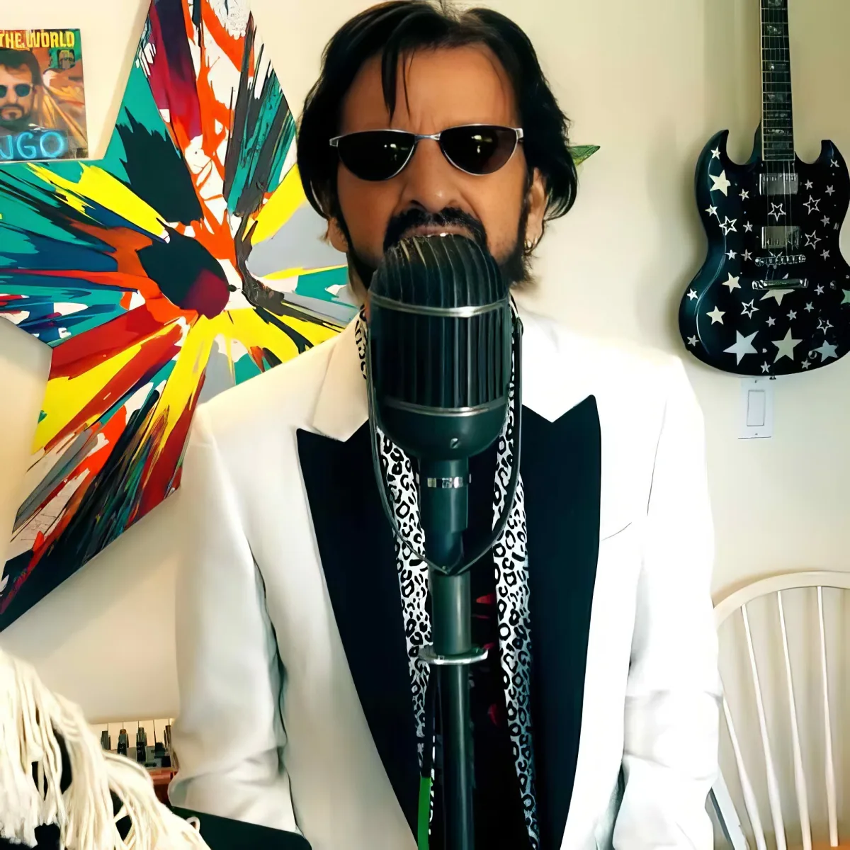 A man in a white suit and sunglasses sings into a microphone, surrounded by colorful art and a guitar.