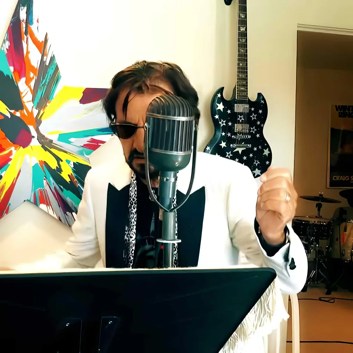 A person in a white suit sings into a microphone in a room with colorful art and a star-patterned guitar.