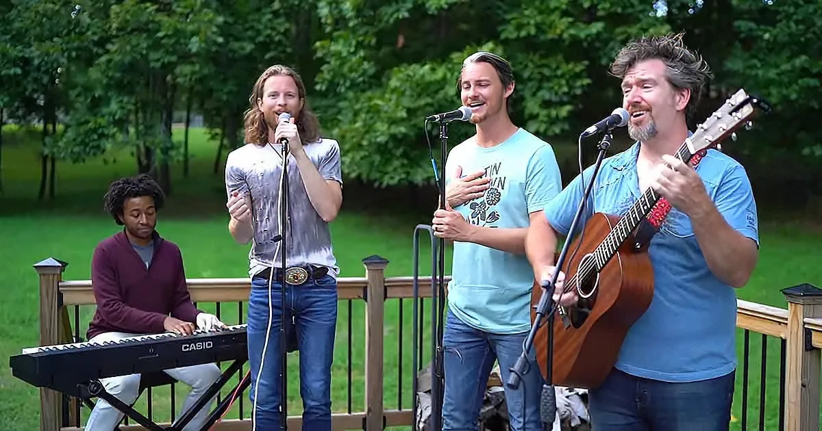 A group of four musicians performs on a deck, with one playing keyboard, two singing, and one playing guitar.