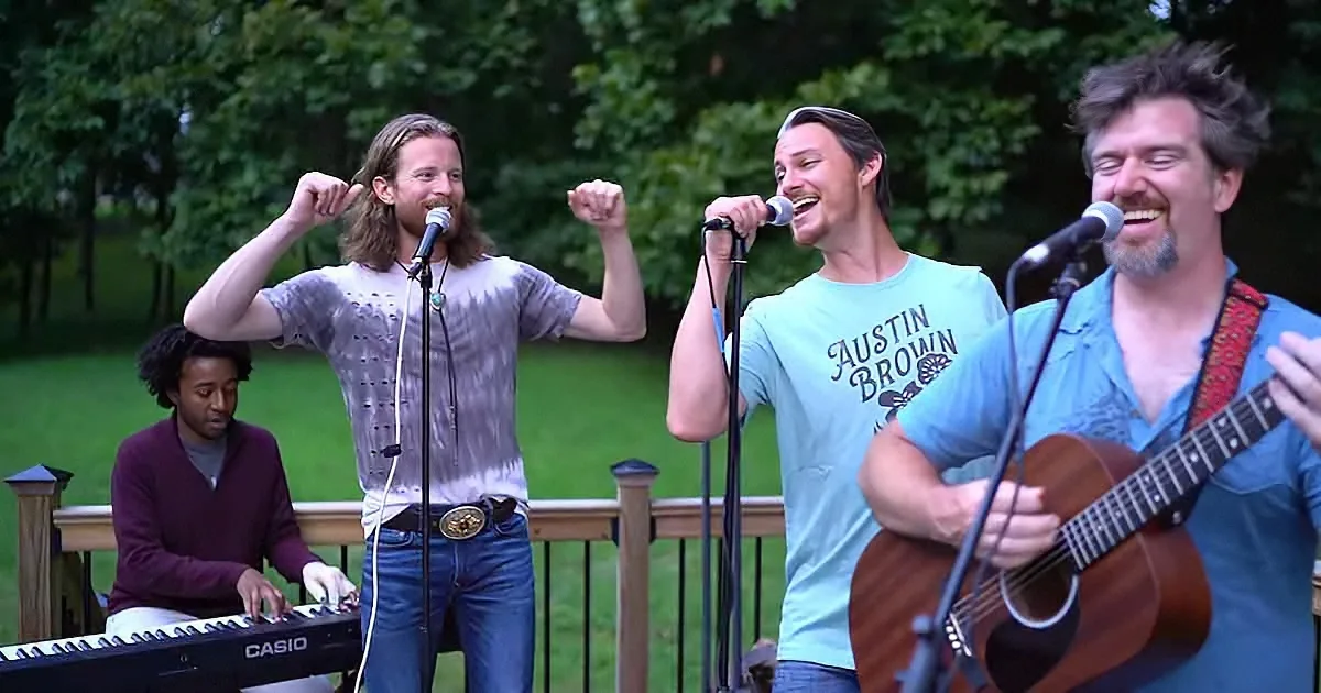 A group of musicians, including a keyboardist, a guitarist, and two singers, perform outdoors on a deck.