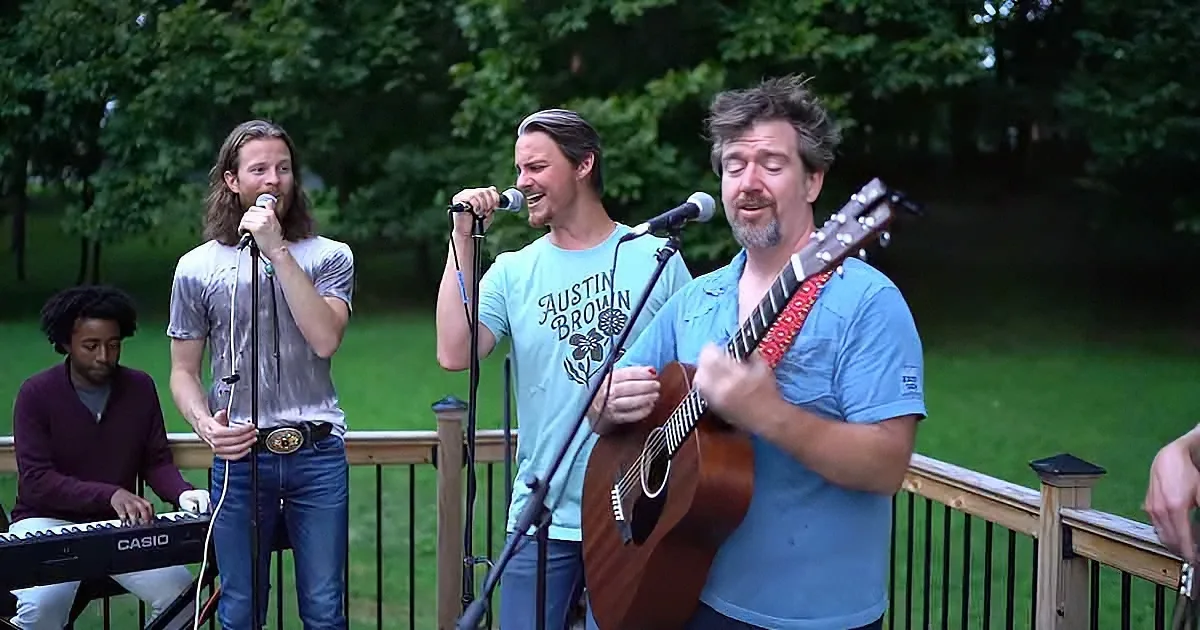 A man in a blue shirt plays guitar while singing, accompanied by others on a deck with a grassy background.