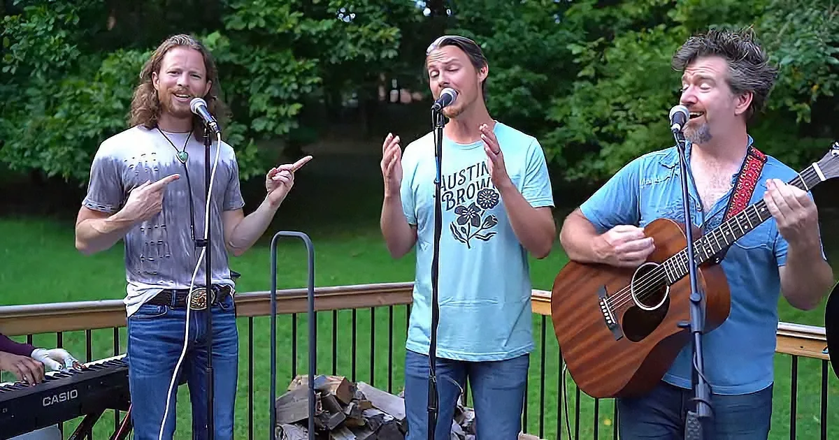 Three men sing and play music outdoors, one playing guitar, another on keyboard, third on vocals.