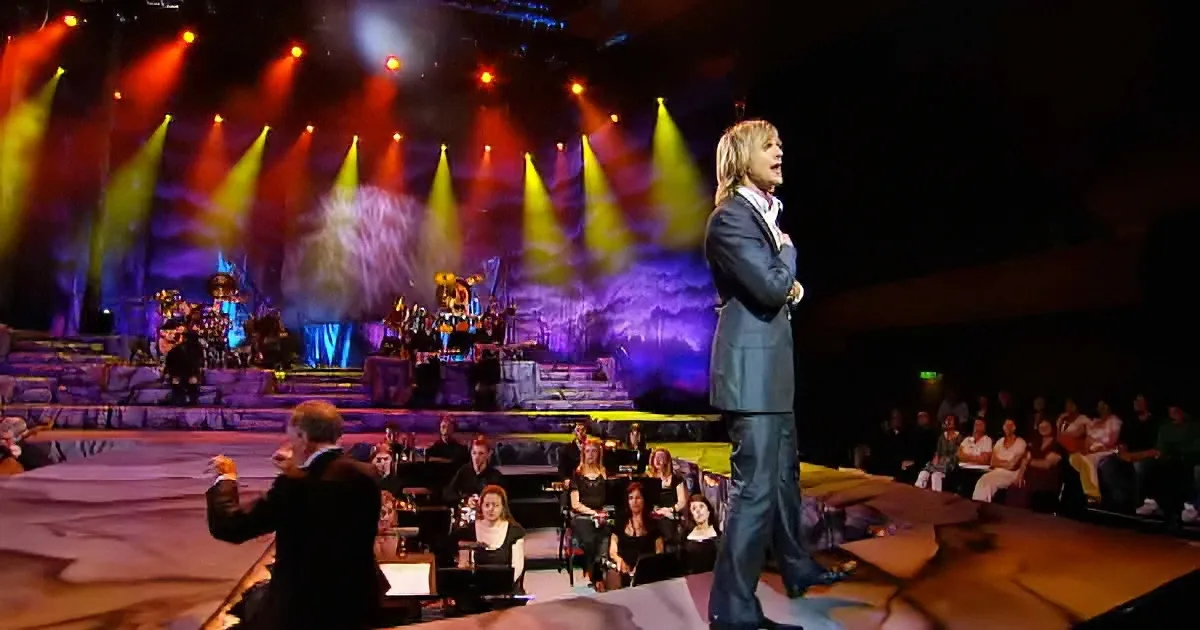 A man in a suit sings on stage with a conductor and orchestra, vibrant lighting.