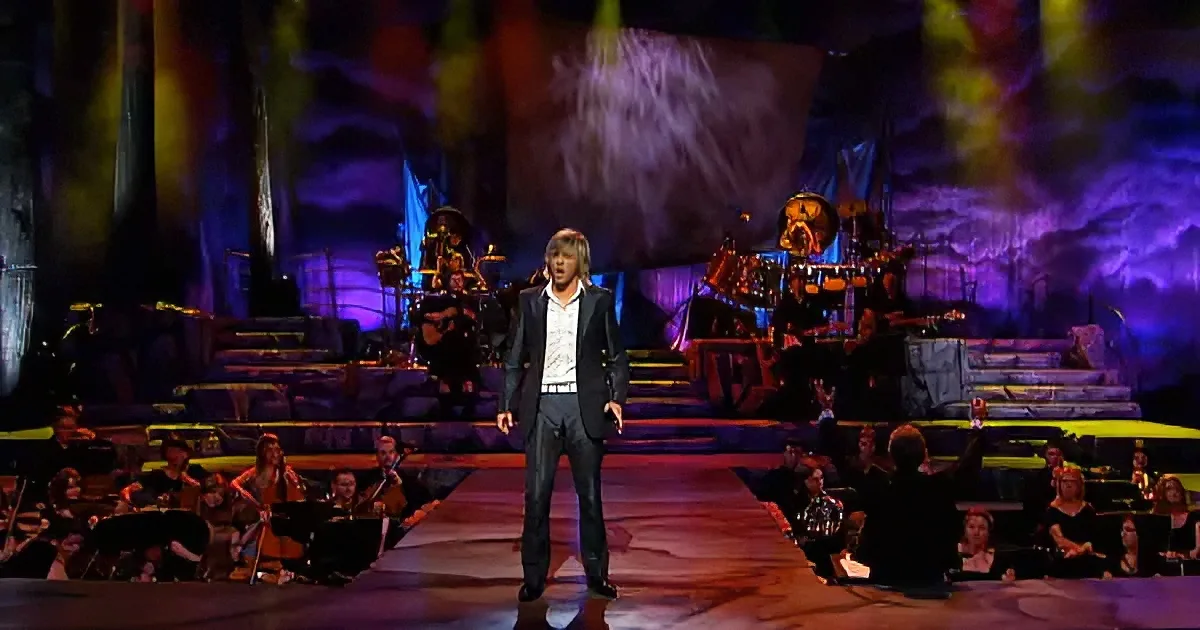 A man in a suit stands on stage with a band and orchestra behind him, illuminated by colorful lights.