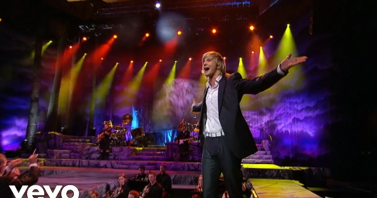A performer in a suit sings passionately on stage with colorful lights and musicians behind.