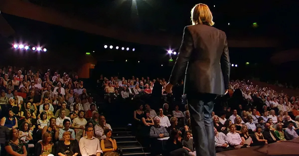 A person with blonde hair stands on stage, facing a large audience in a dimly lit auditorium.