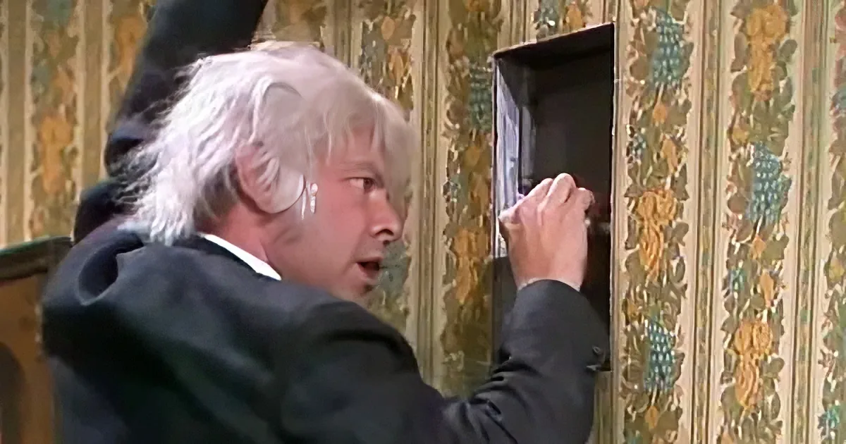 A man in a suit is opening a small cabinet with a surprised expression.