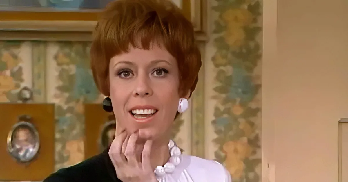 A woman with red hair, wearing a white blouse and black jacket, holds her hand near her mouth in a surprised or shocked...