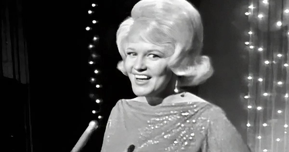 A woman with blonde hair, wearing a sparkly dress, smiles while holding a microphone against a backdrop of twinkling lights.