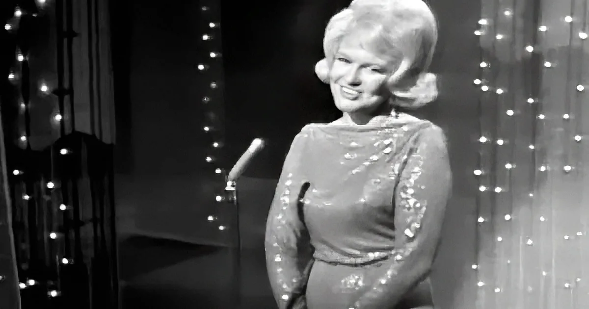 A woman in a sequined dress smiles on stage with twinkling lights in the background.