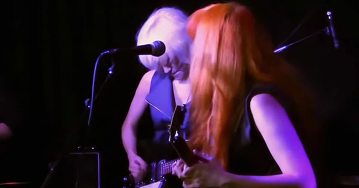 A musician with white hair plays guitar, accompanied by a person with long red hair.