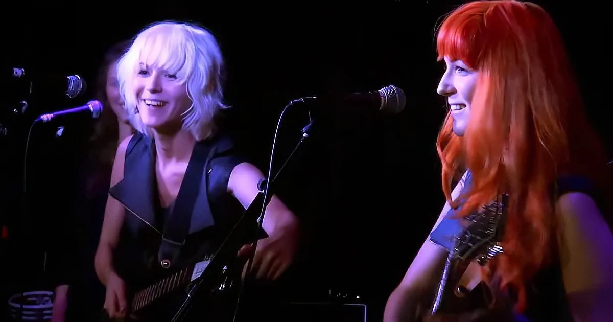 Two women with guitars smile on stage, one with blonde hair, the other with red, under dim lighting.