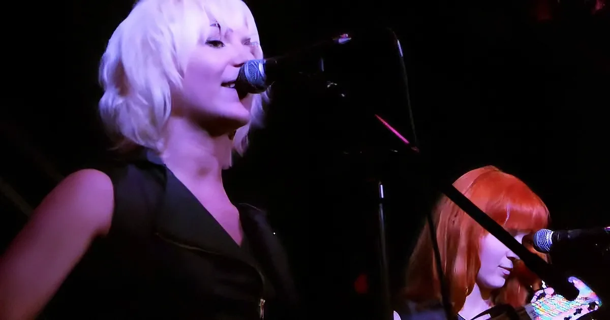 A woman with short white hair sings into a microphone on stage, illuminated by stage lights.