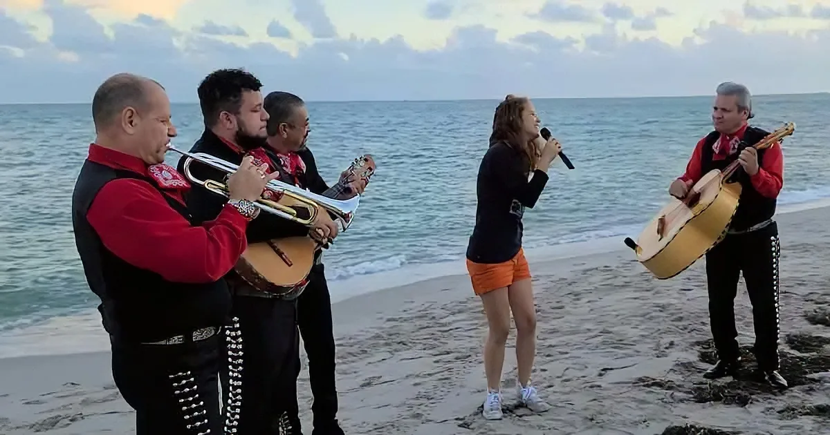 A group of musicians in mariachi attire performs on a beach with a singer.