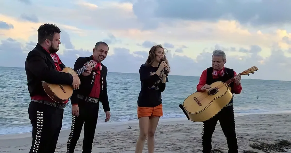 A group of musicians and a singer perform on a beach, playing guitars and singing under a cloudy sky.