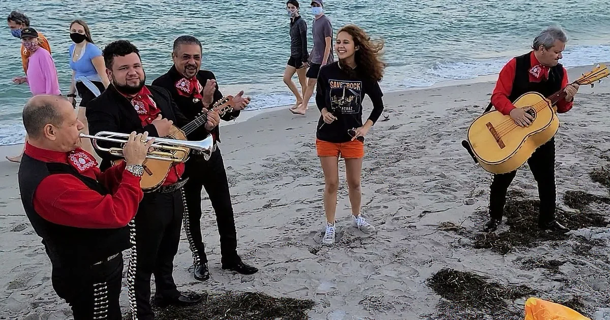 A woman sings on a beach with a mariachi band playing guitars and a trumpet.