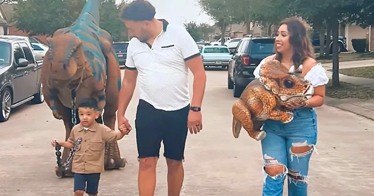 A family walks down a suburban street, the child holding hands with a man, while a woman carries a dinosaur toy and...