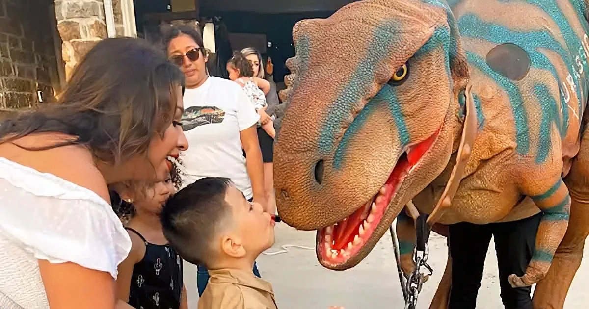 A woman and children interact with a large, realistic T-Rex costume at a party.