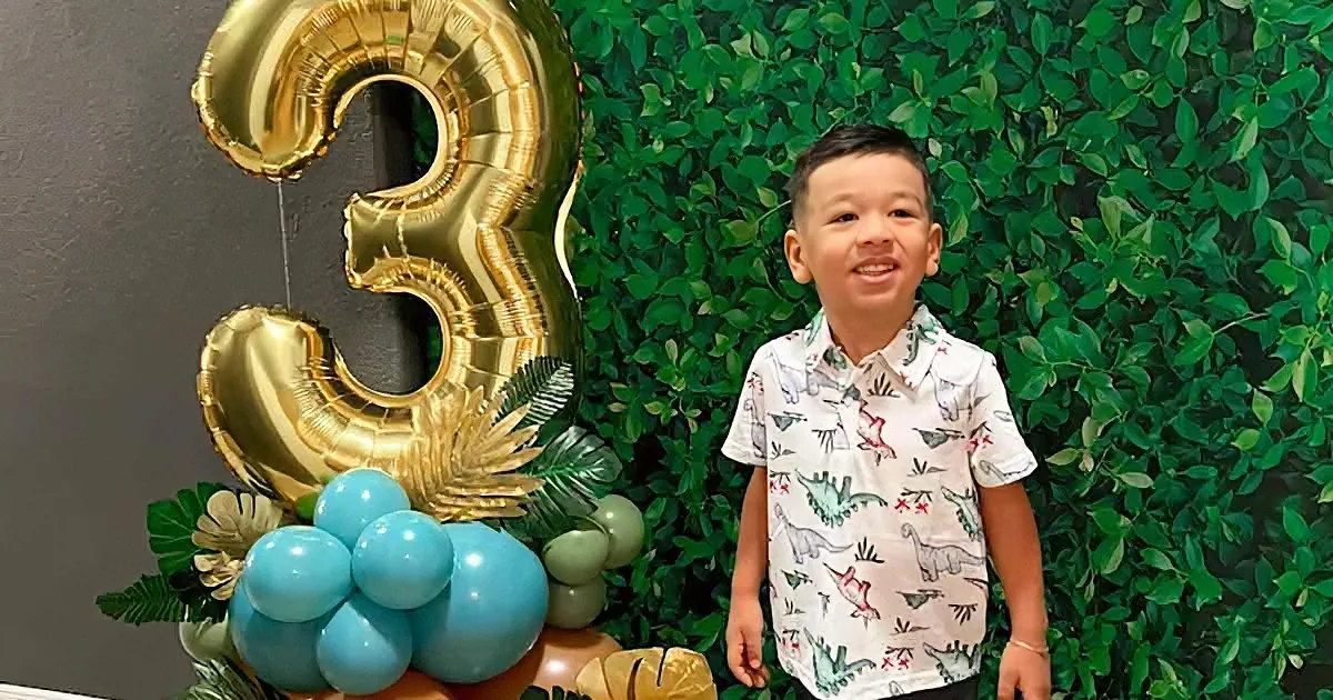 A young boy in a dinosaur shirt smiles in front of a green leafy backdrop with a large gold number 3 balloon.