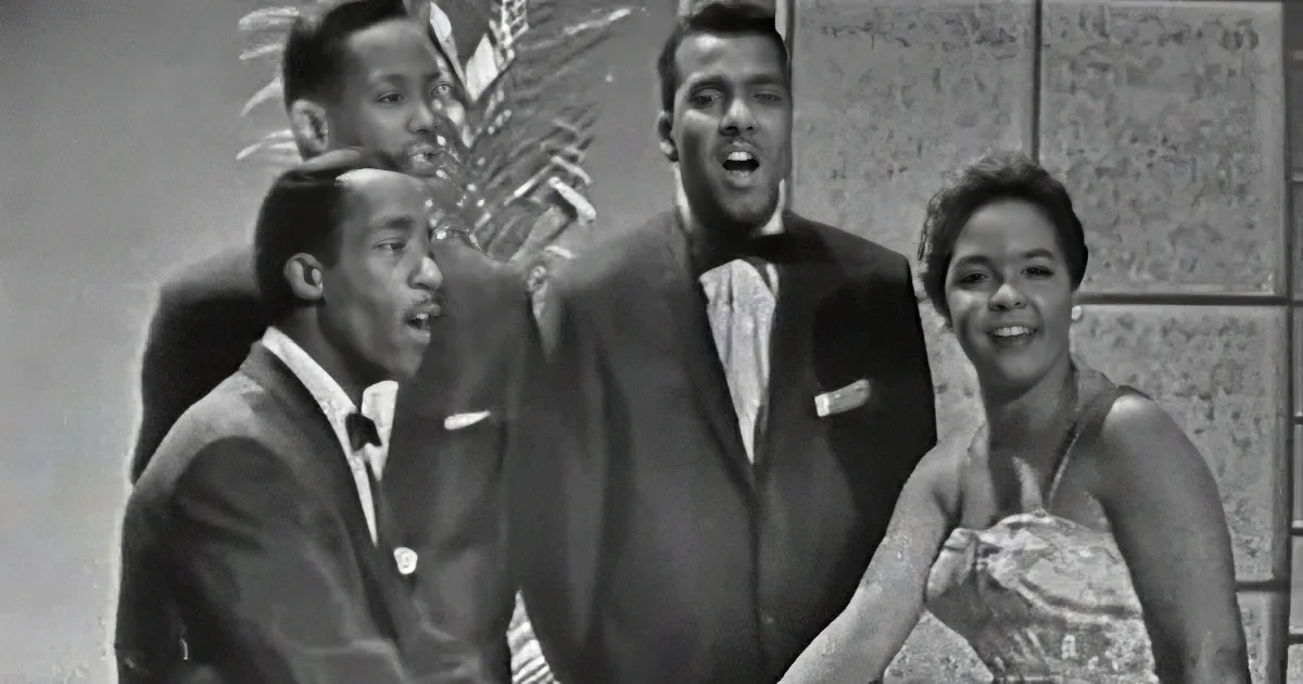 A group of four people, three men in suits and one woman in a dress, appear to be singing or performing.