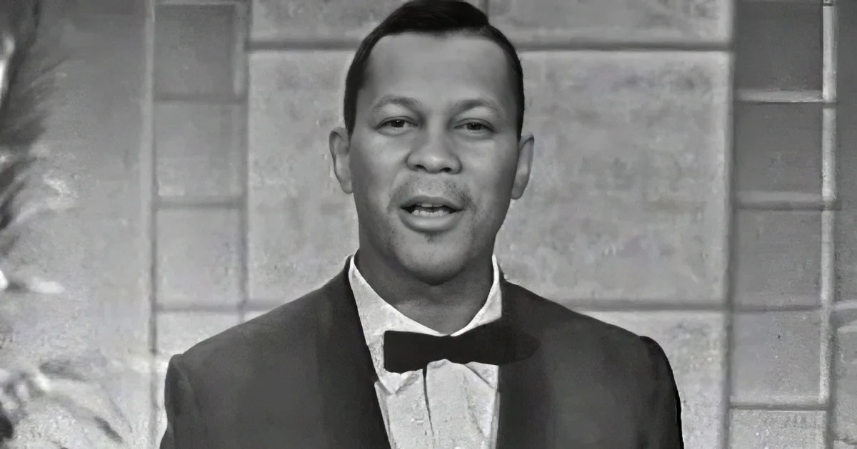 A man in a tuxedo with a bow tie, smiling, against a brick wall backdrop.