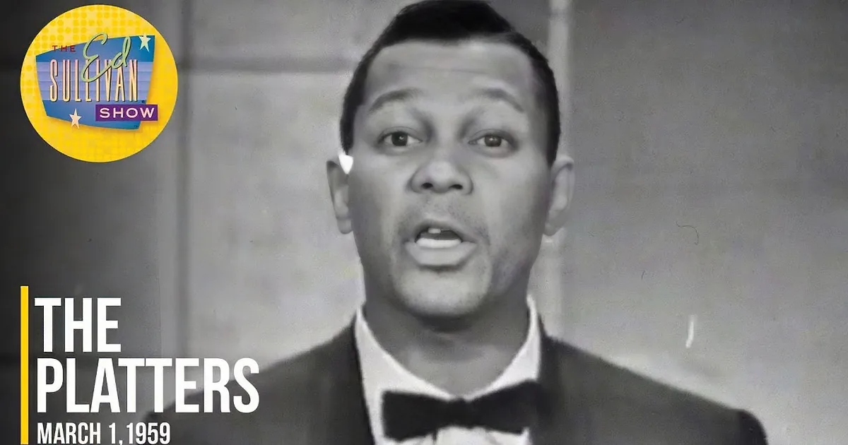 A man in a tuxedo sings in the foreground, with a group of men in suits behind him.