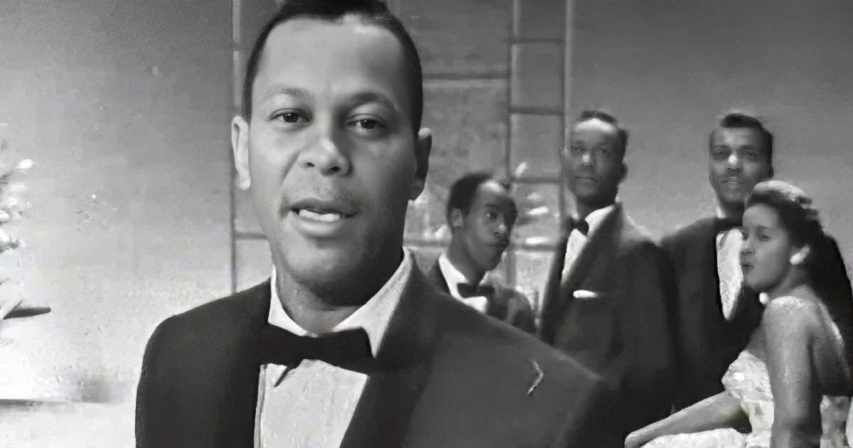 A man in a tuxedo stands in the foreground, singing, with a group of people in the background.