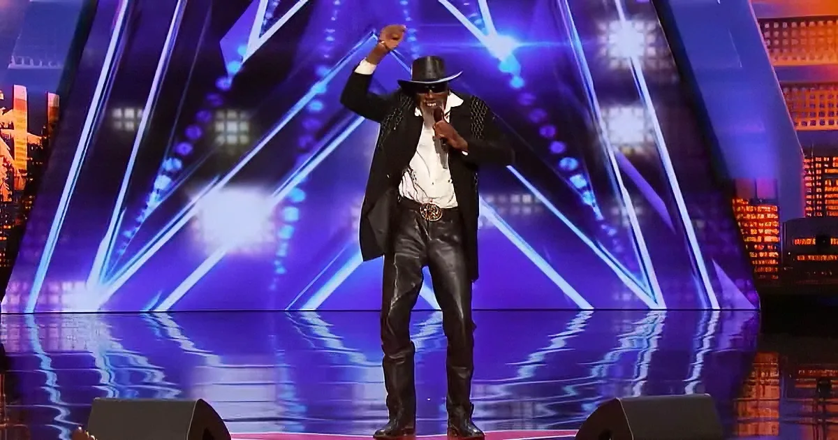 A man in a black hat and leather outfit stands on stage, holding a microphone, with a vibrant blue and purple backdrop.