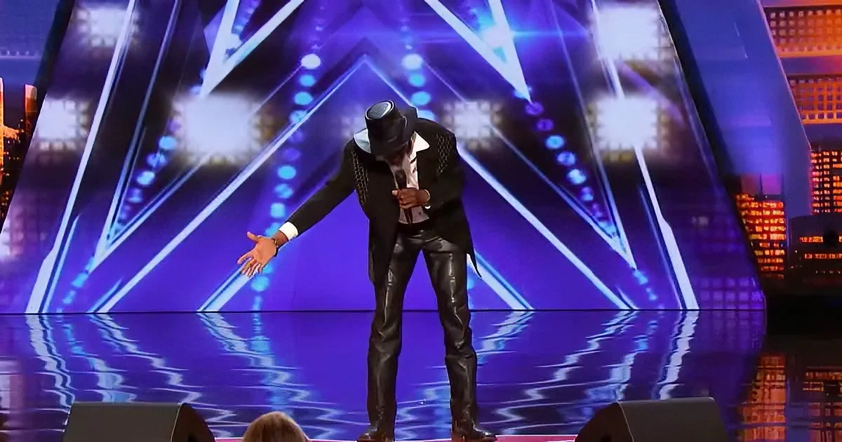 A man in a black suit and hat stands on stage, holding a microphone, with a vibrant purple and blue background.
