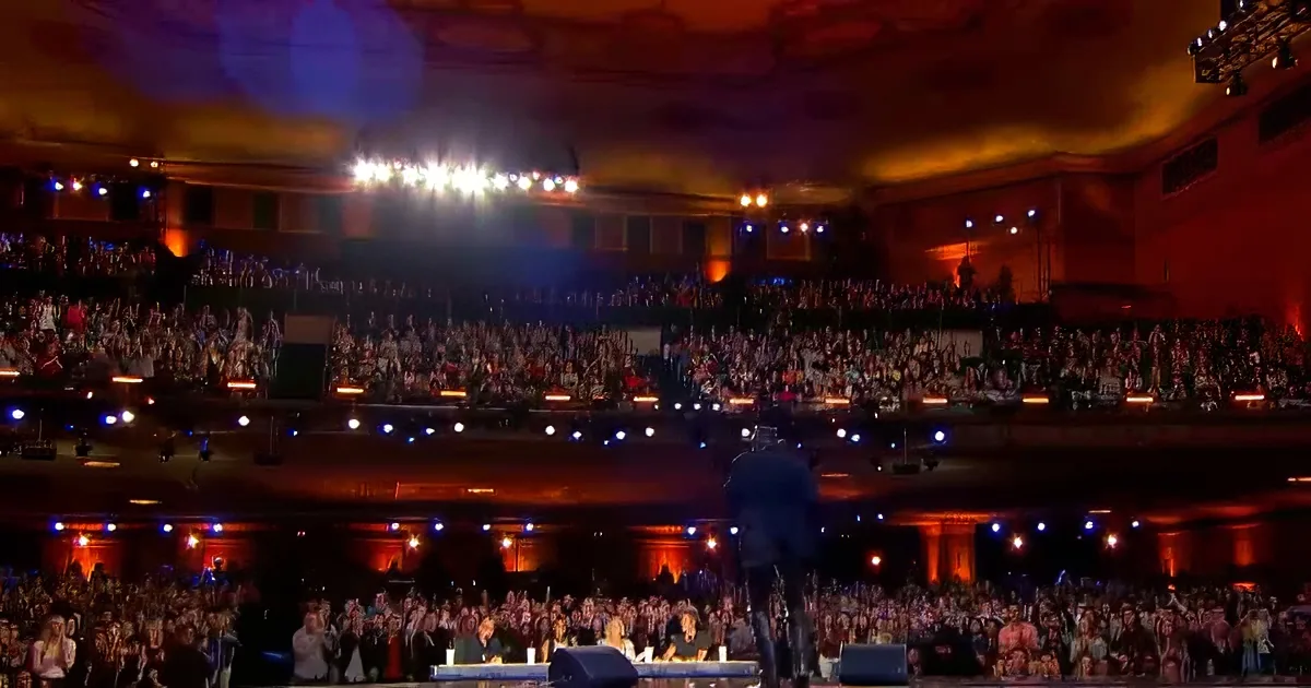 A performer stands on stage in front of a large, enthusiastic audience in a grand theater.