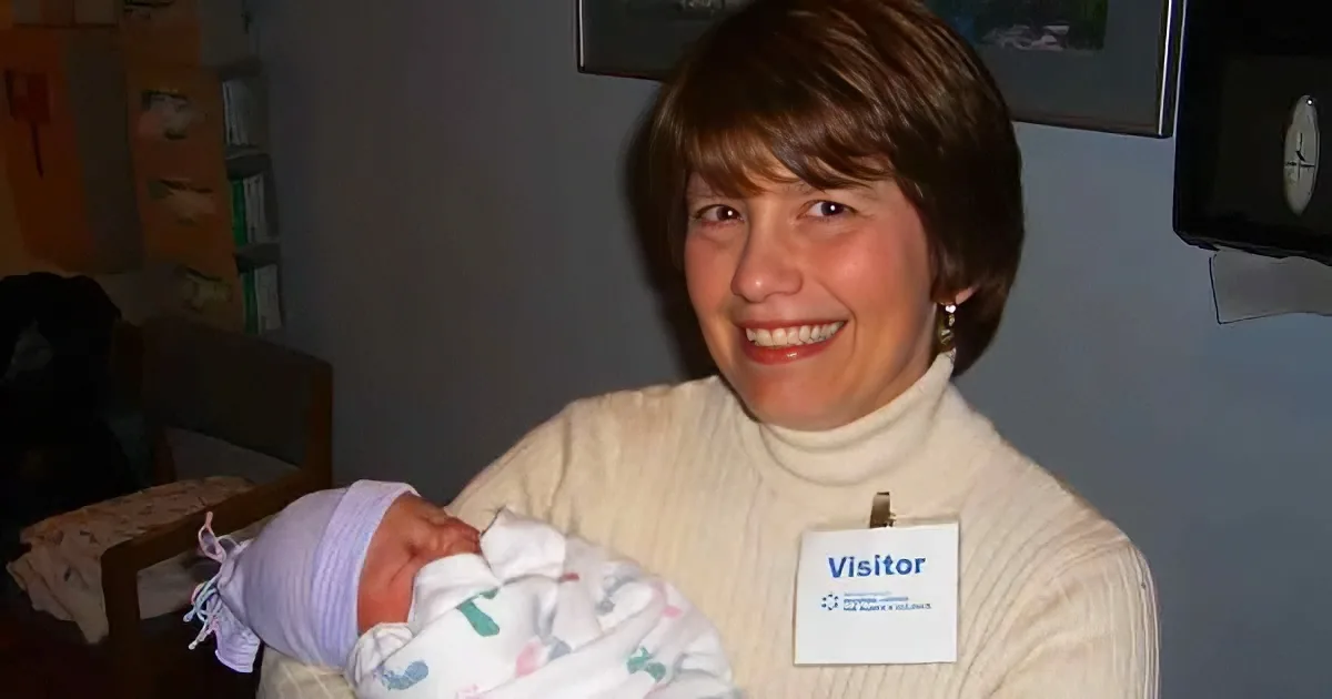 A woman in a white turtleneck holds a newborn baby wrapped in a blanket, smiling.