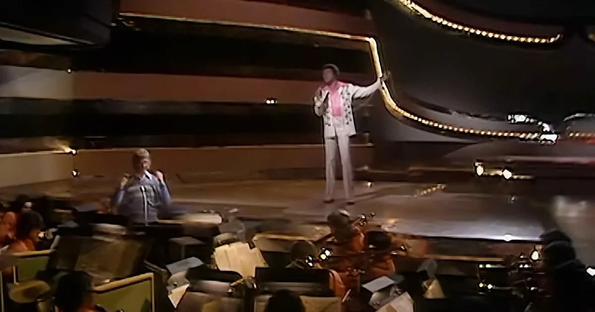A man in a white suit and pink scarf stands on stage, holding a microphone, with an orchestra and audience in the foreground.