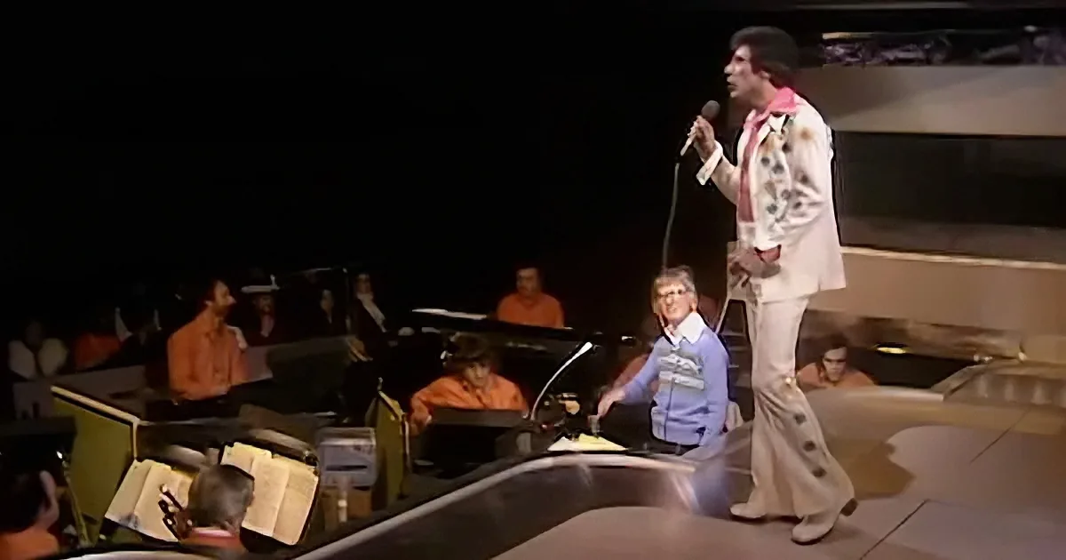 A man in a white suit sings into a microphone on stage with an orchestra behind him.
