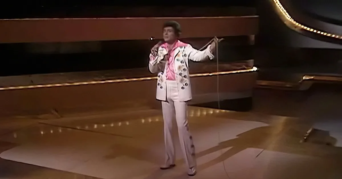 A performer in a white suit with floral patterns sings on stage, holding a microphone.