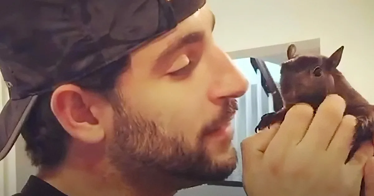 A man with a beard and a black cap holds a small brown animal, possibly a squirrel, in his hands.