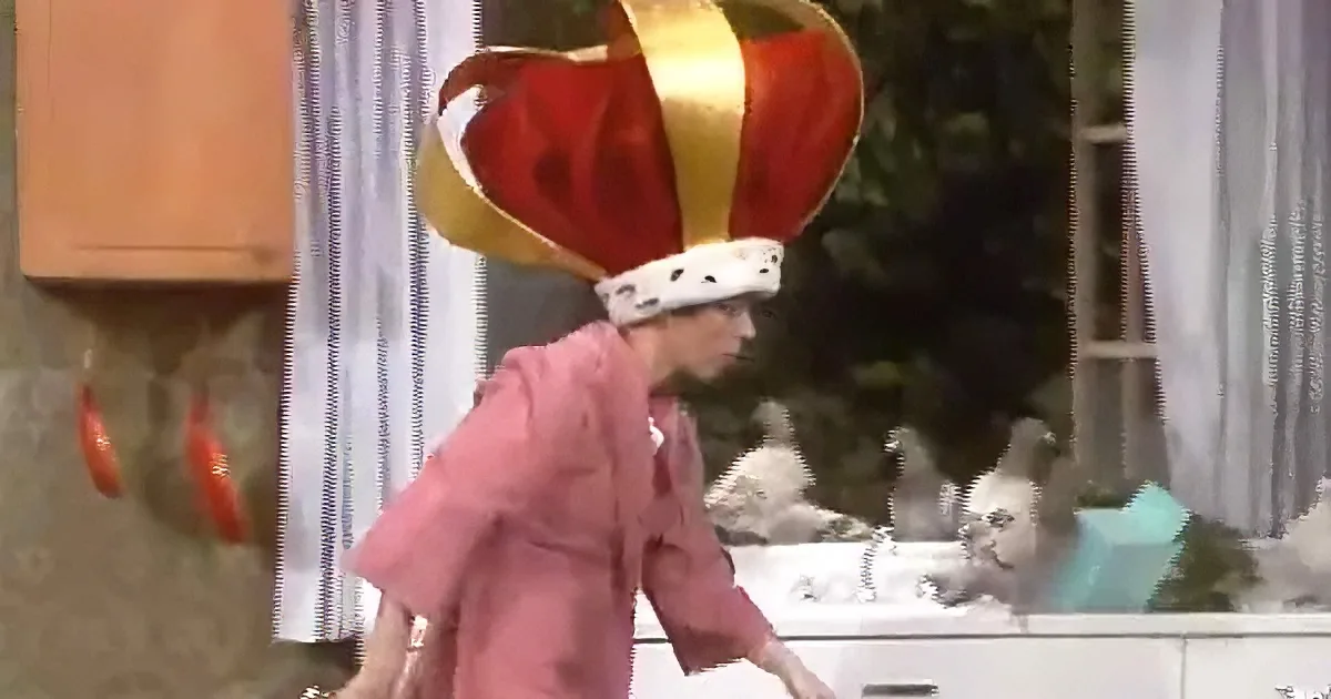 A person in a pink coat and large red crown stands in a room with a fish tank and a window.