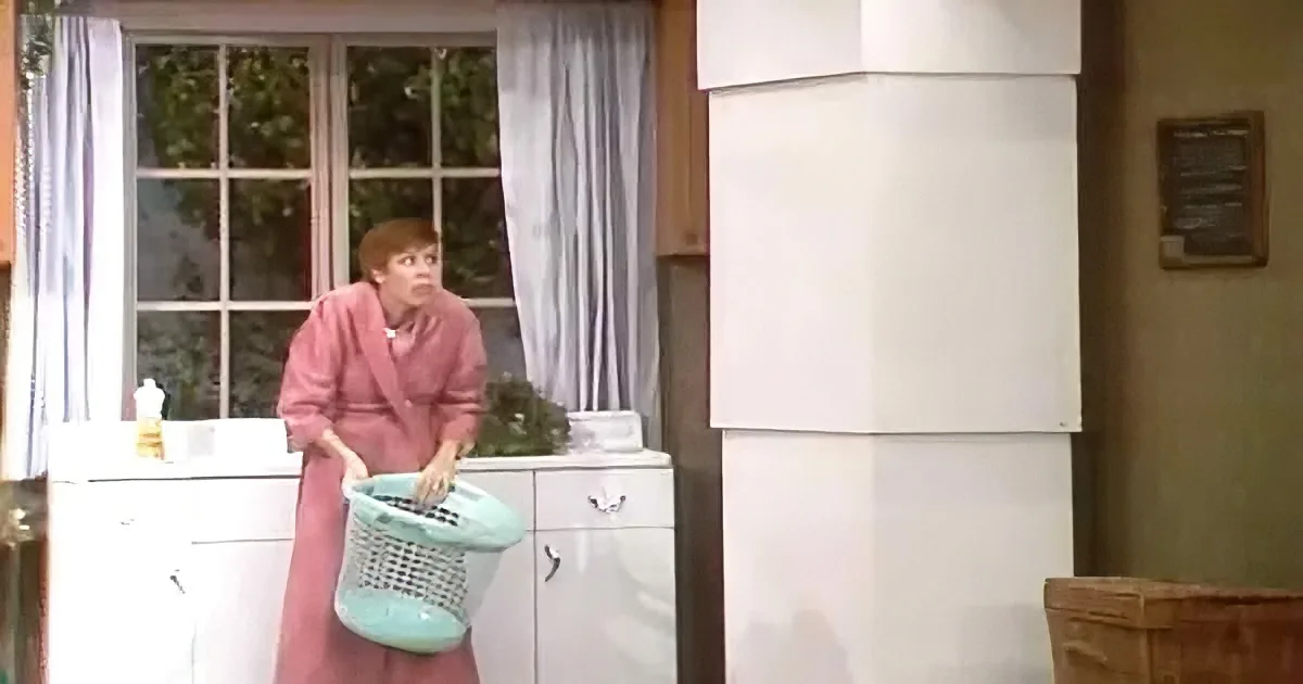 A woman in a pink dress holds a green laundry basket in a room with a white cabinet and window.