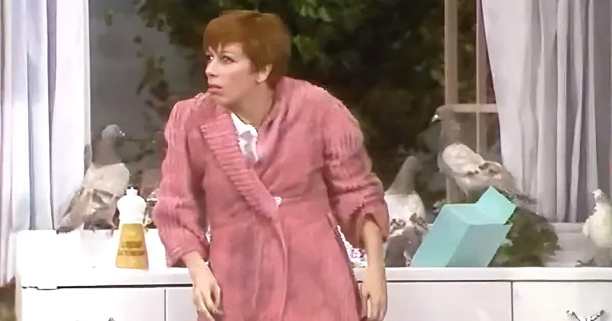 A woman in a pink robe stands in a room with a white dresser, a yellow bottle, and a blue box.