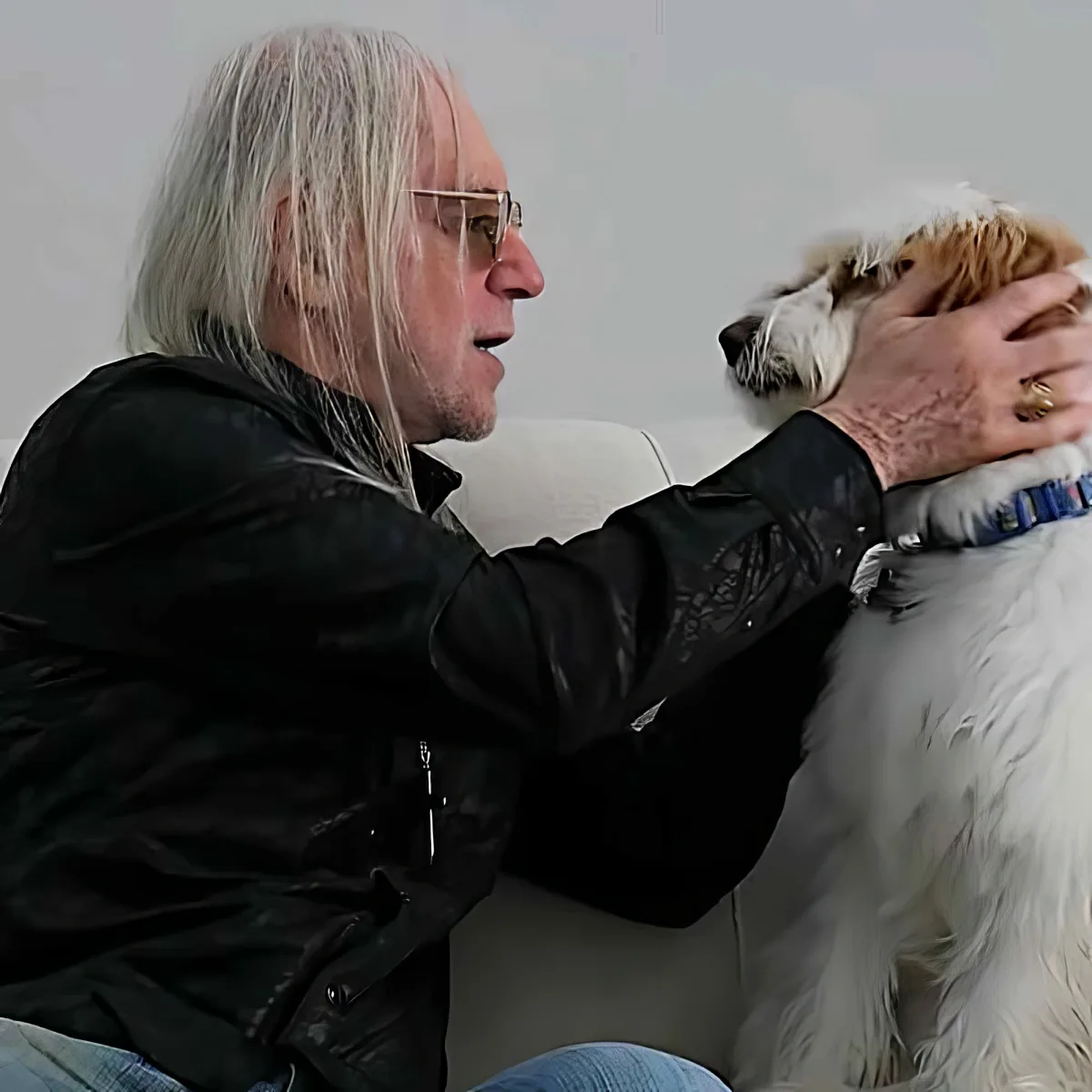 An older man with long white hair and glasses pets a white dog with a blue collar.