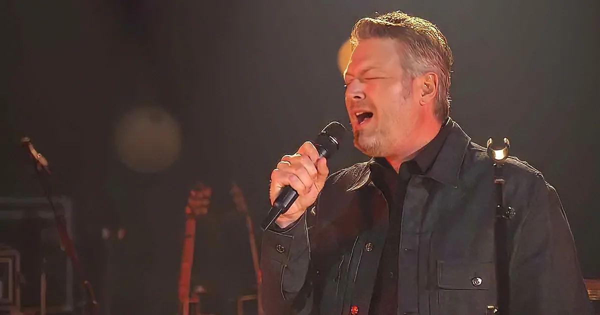 A man in a black jacket sings passionately into a microphone on stage.