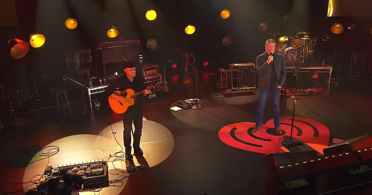 A man sings into a microphone on stage, accompanied by another playing guitar, with a red guitar logo on the floor.