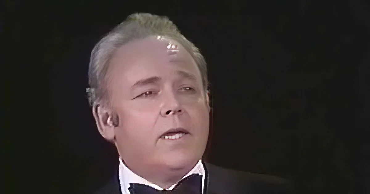 A man in a tuxedo with a bow tie appears to be singing or speaking against a dark background.