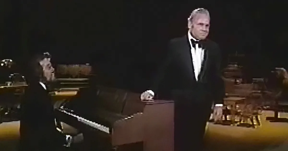 A man in a tuxedo stands beside a seated pianist in a dimly lit stage setting.