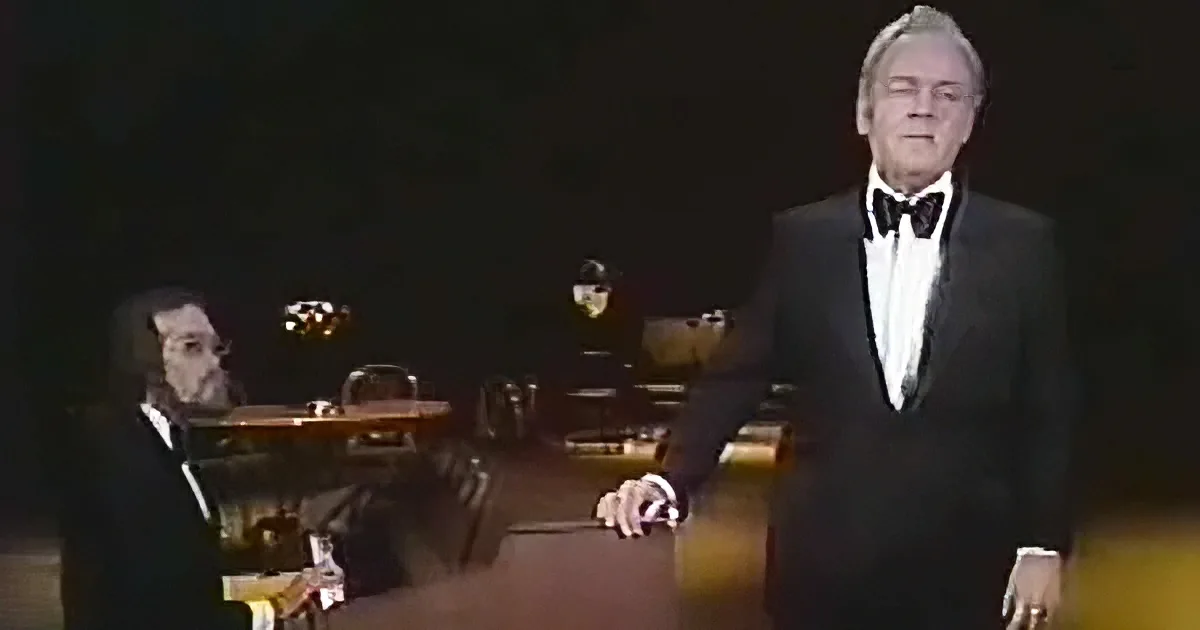 A man in a tuxedo stands in a dimly lit room, with another man seated at a piano in the background.