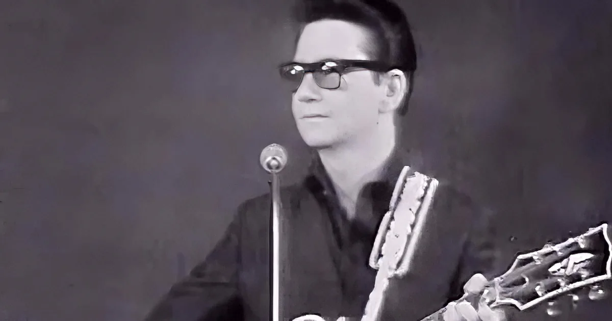 A man in glasses plays a guitar, wearing a dark shirt, in front of a microphone.