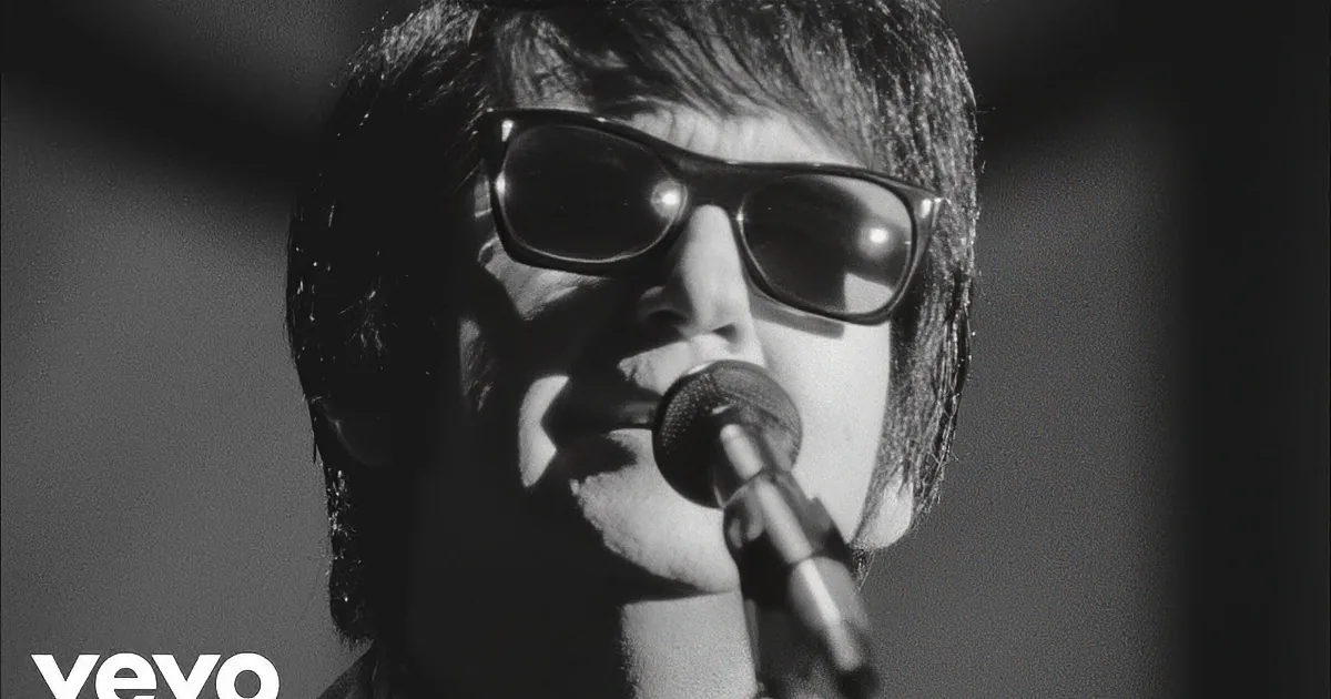 A man in a suit and sunglasses holds a guitar, standing against a dark background.